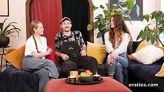 Two women kiss naked while man watches from behind plants
